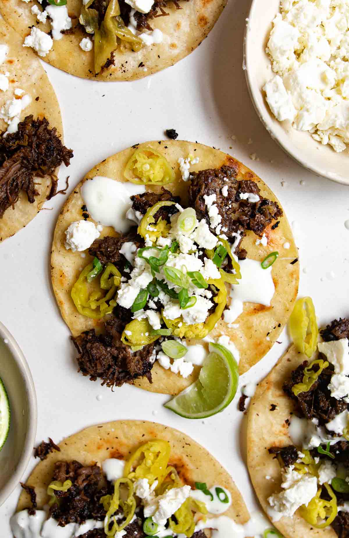 Five Mississippi Pot Roast Tacos topped with sour cream, cotija cheese, green onions and pepperoncini on a table.