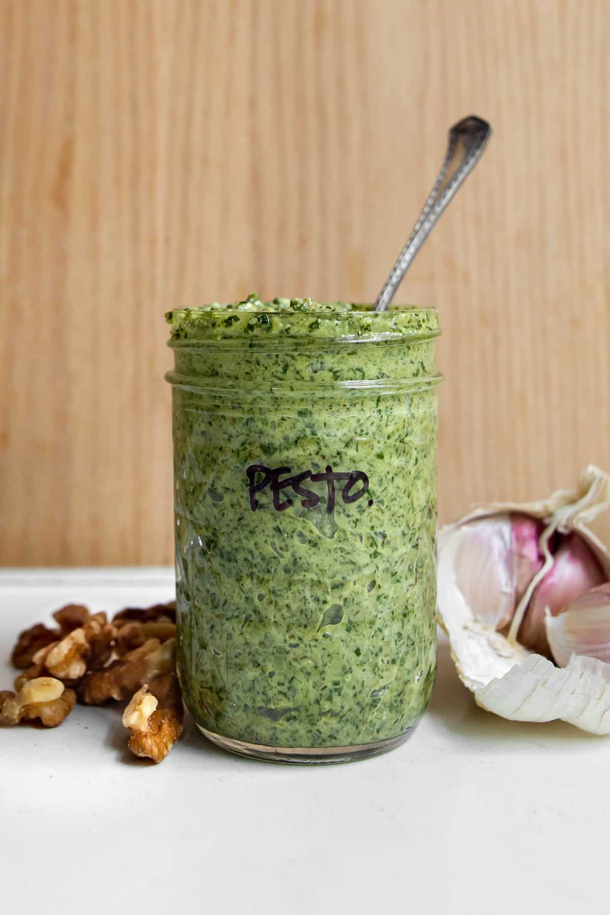side shot of jar with pesto and a spoon with walnuts and garlic around it.