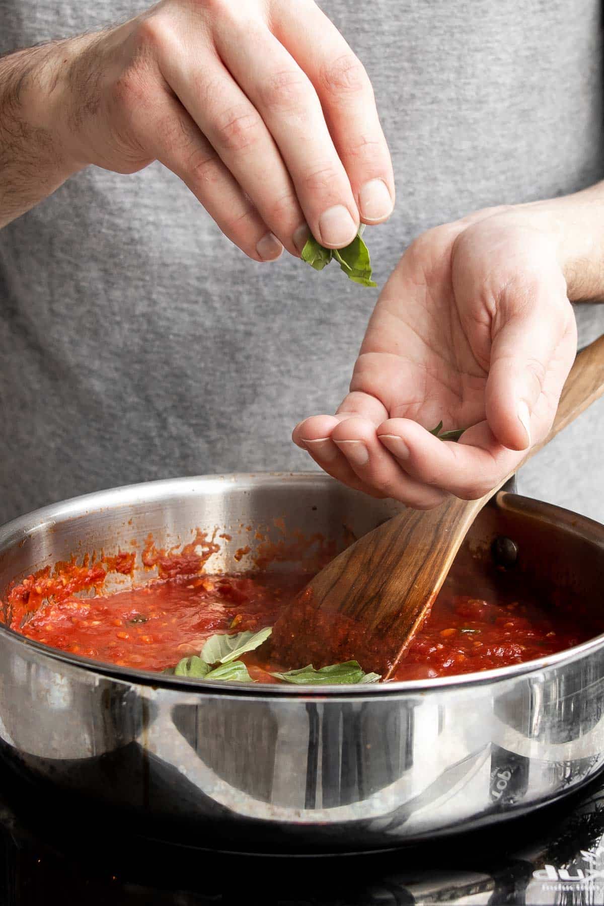 hand adding basil to marinara sauce in a pan.