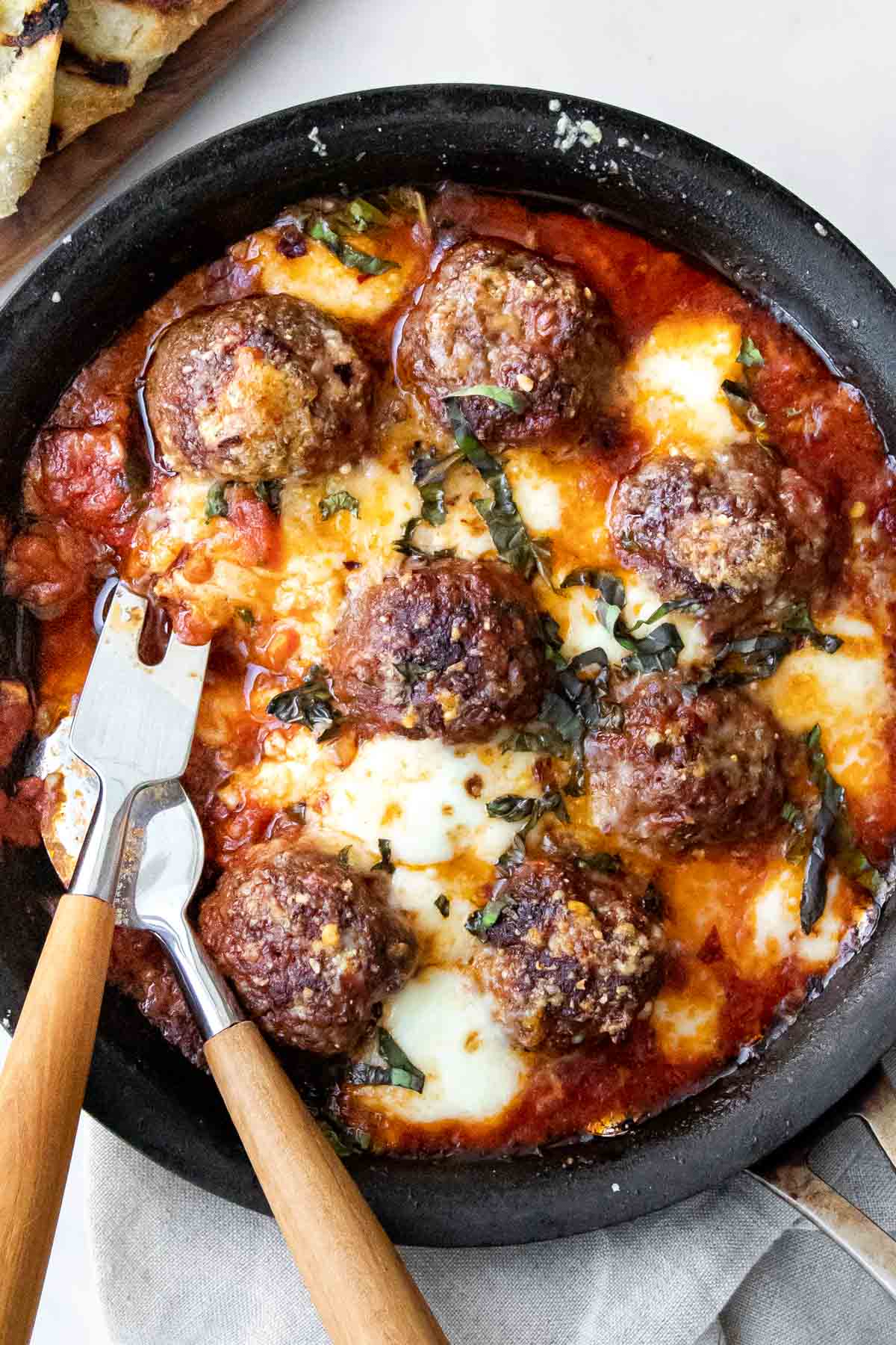spicy meatballs with marinara sauce, baked mozzarella and fresh basil in a cast-iron skillet with serving utensils.