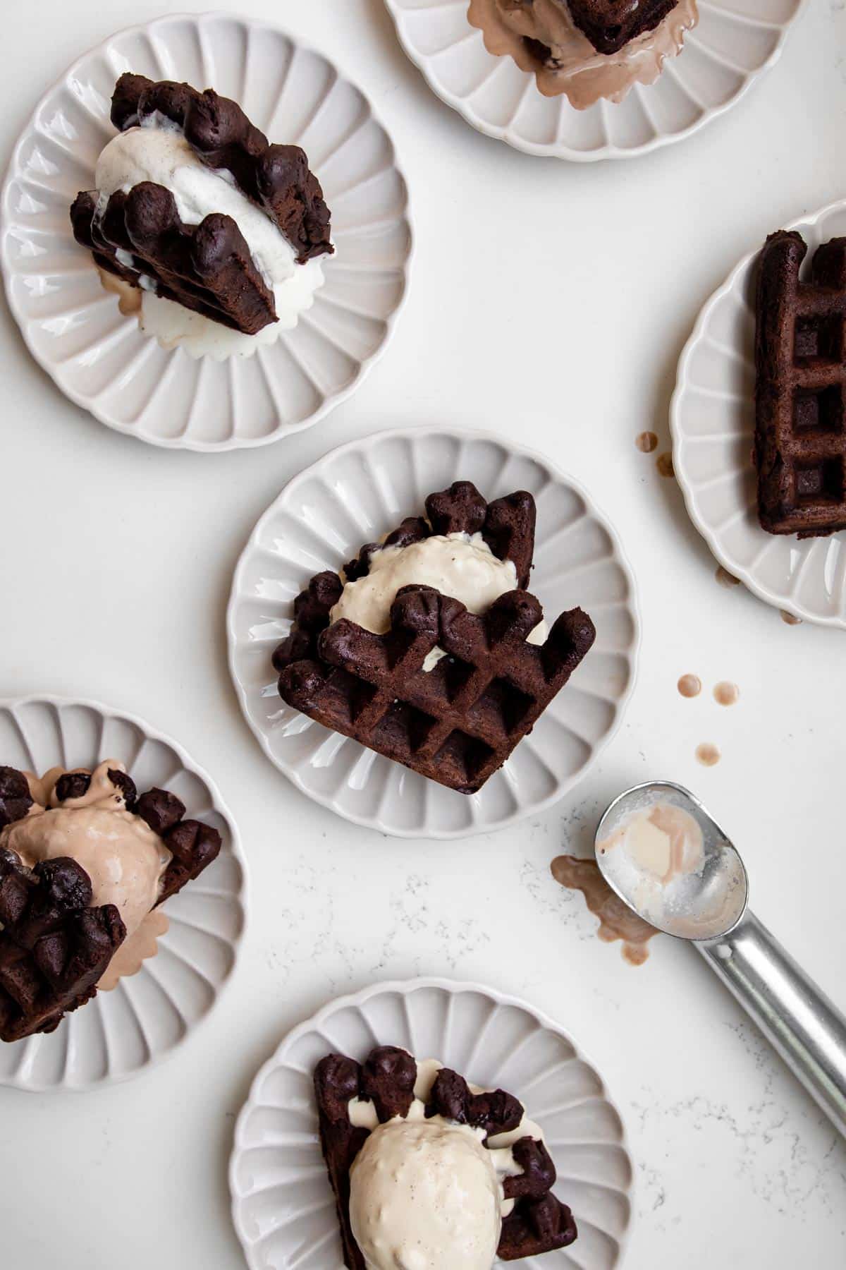 Six brownie waffle ice cream sandwiches on plates with an ice cream scoop.
