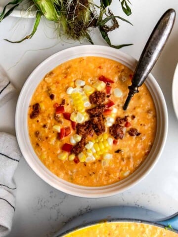 corn chowder with sausage in a bowl with a spoon with corn husks and pot of soup around it.