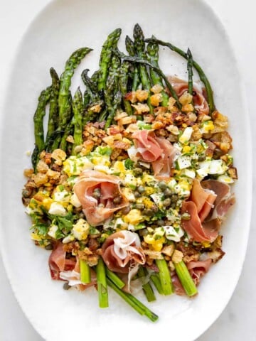asparagus salad topped with boiled eggs, breadcrumbs, capers and prosciutto on a platter.