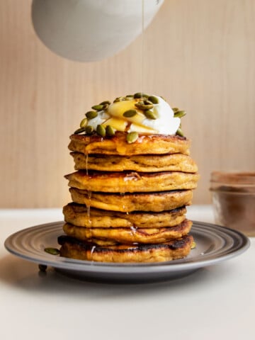 a stack of butternut squash pancakes on a plate topped with yogurt, pumpkin seeds and maple syrup.