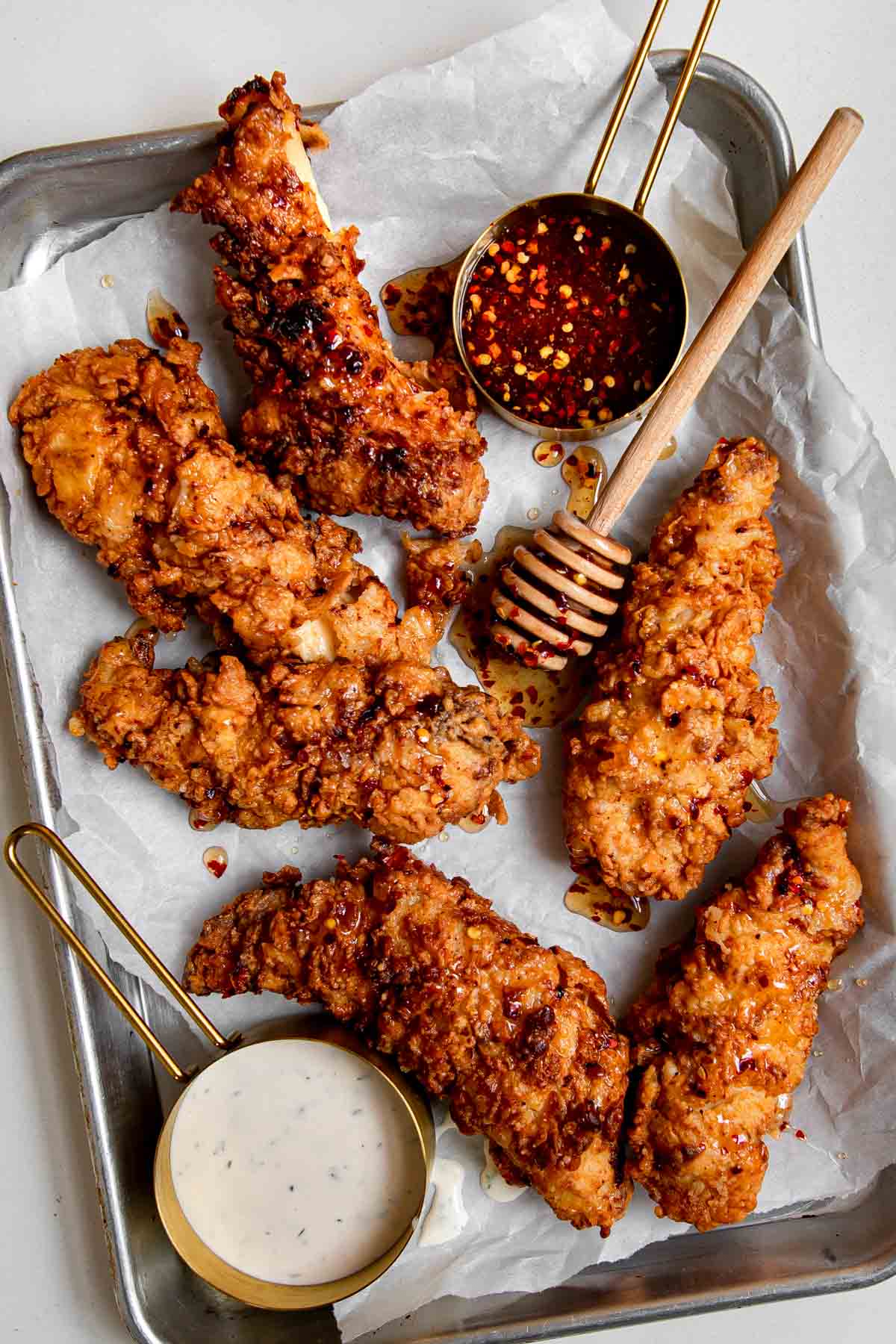 Hot honey tenders with honey dipper, hot honey sauce, and ranch on a parchment lined baking sheet.